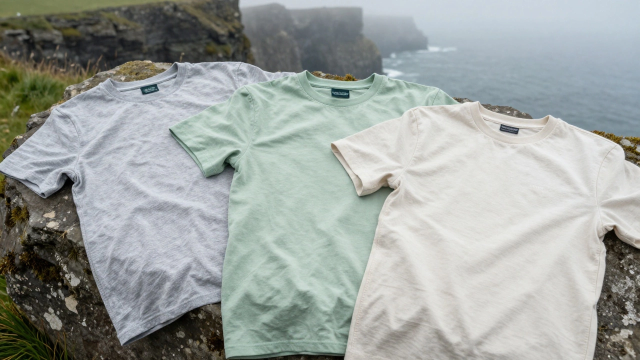 Three Irish-made cotton tees in neutral tones laid on mossy rock near Cliffs of Moher, with mist rolling in.